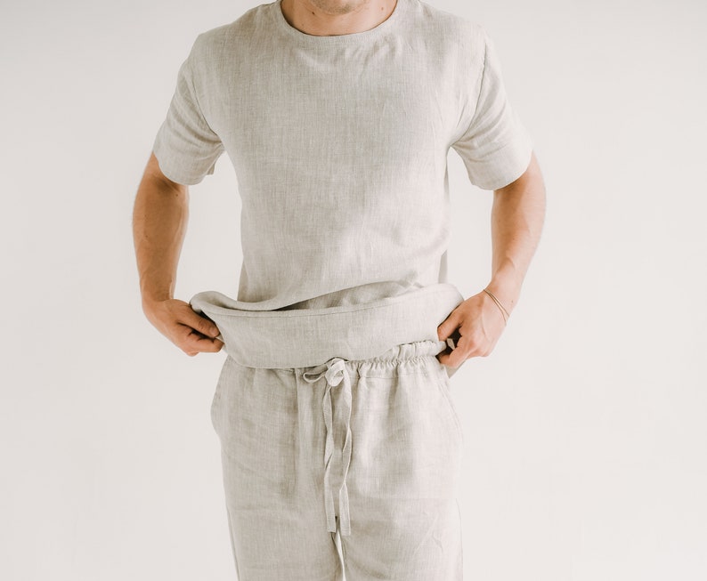 May include: A light grey linen two-piece set. The set includes a short-sleeved crew neck shirt and drawstring trousers. The fabric has a textured appearance. The model is wearing the set. The background is plain white.