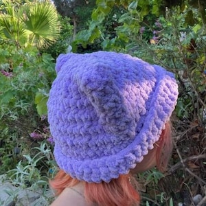 May include: A soft, purple crocheted beanie hat with a fluffy texture. The hat is worn on a person's head with pink hair.