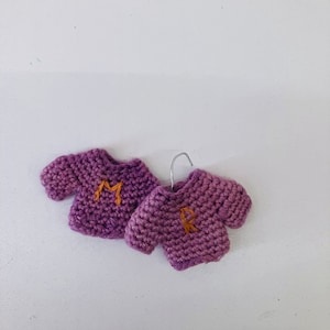 May include: Two miniature purple crocheted sweaters with the letters "M" and "R" embroidered in orange yarn.