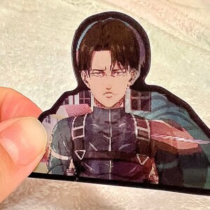 以下が含まれることがあります： アニメ「進撃の巨人」のキャラクターが描かれたステッカー。キャラクターは茶色の髪と緑色の目をした若い男性。彼は赤と黒のベルトが付いた濃い灰色の制服を着ている。彼は右手に剣を持ち、決意に満ちた表情をしている。