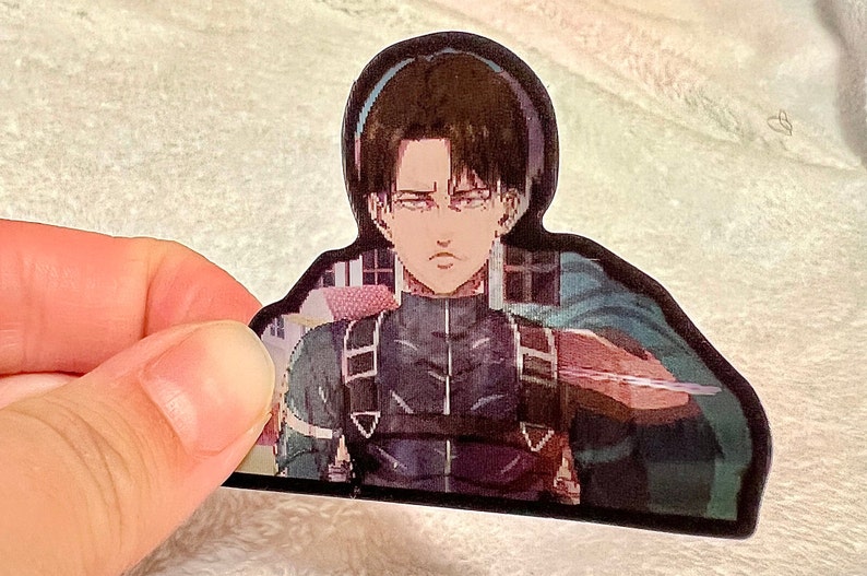 以下が含まれることがあります： アニメ「進撃の巨人」のキャラクターが描かれたステッカー。キャラクターは茶色の髪と緑色の目をした若い男性。彼は赤と黒のベルトが付いた濃い灰色の制服を着ている。彼は右手に剣を持ち、決意に満ちた表情をしている。