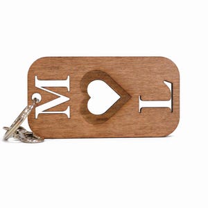 Pode incluir: Um porta-chaves retangular de madeira com cantos arredondados apresentando um recorte da letra M, uma forma de coração e a letra L. O grão da madeira castanha natural é visível e um anel de metal prateado está preso ao lado esquerdo.