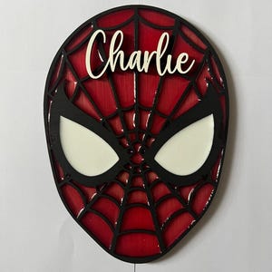 Puede incluir: Una placa de pared de madera en capas con forma de máscara de superhéroe con fondo rojo y un patrón de telaraña negro. La máscara presenta dos grandes aberturas blancas para los ojos y el nombre Charlie en letras cursivas blancas.