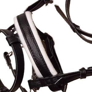 May include: A black leather harness featuring white padded lining along the central strap. The harness includes multiple adjustable straps with metal buckles and loops, designed with a textured finish on the black leather components throughout.