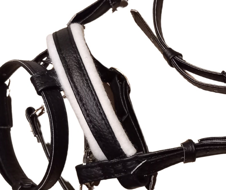 May include: A black leather harness featuring white padded lining along the central strap. The harness includes multiple adjustable straps with metal buckles and loops, designed with a textured finish on the black leather components throughout.