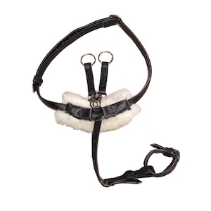Peut inclure: Un harnais en cuir noir doté d'une pièce de poitrine rembourrée en fausse fourrure blanche, de sangles réglables avec boucles métalliques, de deux anneaux en métal argenté et d'une attache circulaire reliée par une sangle verticale centrale.