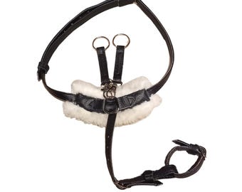 May include: A black leather harness featuring a white faux fur padded chest piece, adjustable straps with metal buckles, two silver-toned metal rings at the top, and a smaller circular strap attachment connected by a central vertical leather strap.