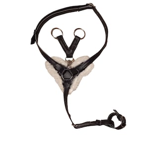 May include: A black leather harness featuring a heart-shaped centre piece lined with white faux fur. The harness includes adjustable straps with metal buckles, silver-toned metal rings, and a central clasp connecting the various strap segments.