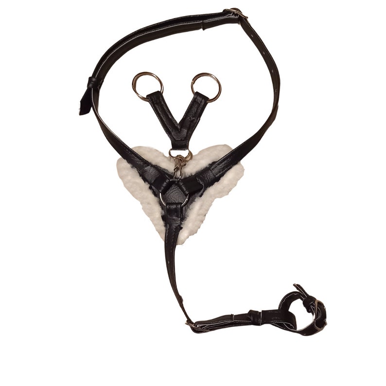 May include: A black leather harness featuring a heart-shaped centre piece lined with white faux fur. The harness includes adjustable straps with metal buckles, silver-toned metal rings, and a central clasp connecting the various strap segments.