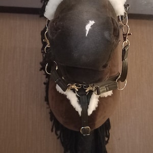 May include: A brown hobby horse with a white blaze on its nose, featuring a black mane and a black bridle with white faux fur trim. The bridle includes metal rings and clips, and the horse is mounted on a wooden stick handle for support.