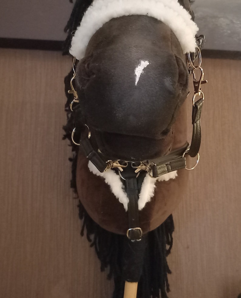 May include: A brown hobby horse with a white blaze on its nose, featuring a black mane and a black bridle with white faux fur trim. The bridle includes metal rings and clips, and the horse is mounted on a wooden stick handle for support.