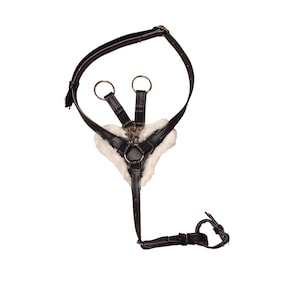 May include: A black leather harness featuring a white faux fur lining on the chest piece. The harness includes adjustable straps, silver-toned metal rings, and a central metal clasp, designed with a Y-shaped configuration for secure attachment.