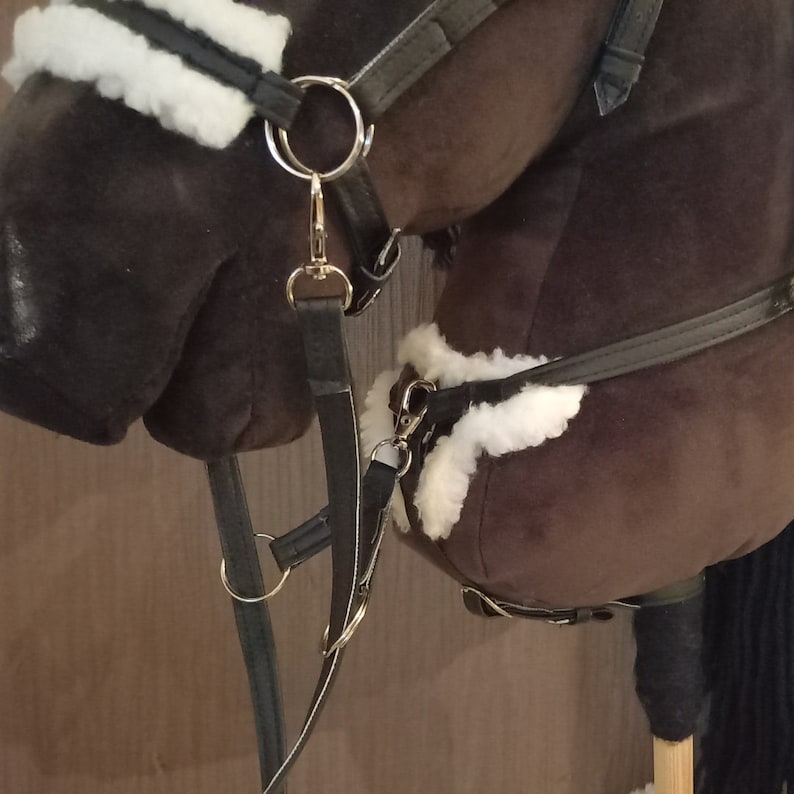 May include: A dark brown plush hobby horse head featuring a black bridle with white faux sheepskin padding on the noseband and browband. The bridle includes silver-toned metal rings, buckles, and clips, with black leather-like straps attached.