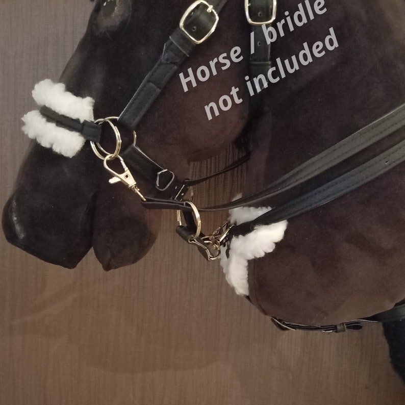May include: A black leather headcollar featuring white faux sheepskin padding on the noseband and throat latch, secured with gold-toned metal rings and clips. The text "Horse / bridle not included" is printed in white on the dark brown plush horse head.