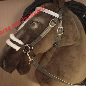 Puede incluir: Una brida negra con una frontalera de purpurina plateada, acolchado de piel sint&eacute;tica blanca en la muserola y hebillas met&aacute;licas. La brida se muestra sobre una cabeza de caballo de peluche marr&oacute;n con crines de lana negra.