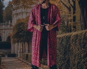 Puede incluir: Una bata tipo kimono larga y abierta por delante, con una base de color magenta intenso y un patrón geométrico repetitivo en dorado y azul verdoso. La prenda tiene mangas anchas de tres cuartos y un borde magenta oscuro sólido.