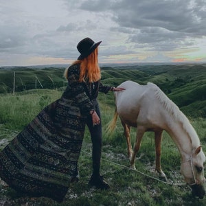 Puede incluir: Una mujer con un abrigo largo estampado y un sombrero negro se encuentra en un campo con un caballo blanco. El caballo est&aacute; pastando en la hierba verde. El cielo est&aacute; nublado y el sol se est&aacute; poniendo en la distancia.