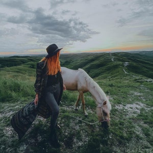Puede incluir: Una mujer con un sombrero negro y un abrigo largo estampado se encuentra con un caballo blanco en un campo de hierba. El cielo est&aacute; nublado y el sol se pone en la distancia.