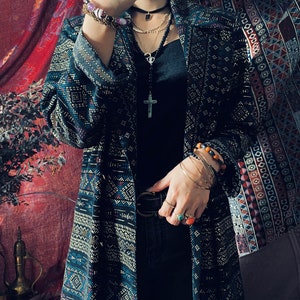 Puede incluir: Una larga chaqueta de punto negra y azul con detalles dorados. La chaqueta tiene cuello y cintur&oacute;n. La persona lleva un top negro sin mangas, vaqueros negros y varias pulseras y collares.