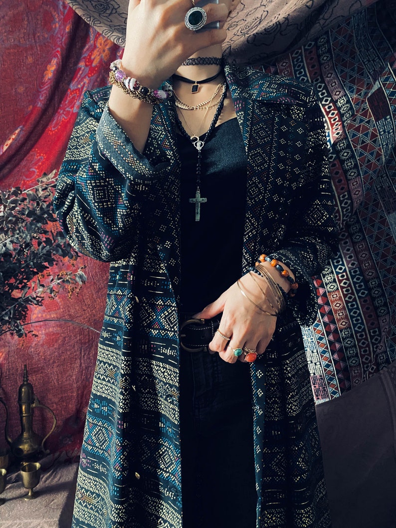 Puede incluir: Una larga chaqueta de punto negra y azul con detalles dorados. La chaqueta tiene cuello y cintur&oacute;n. La persona lleva un top negro sin mangas, vaqueros negros y varias pulseras y collares.