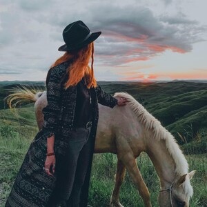 Puede incluir: Una mujer con un sombrero negro y un largo abrigo negro estampado se encuentra junto a un caballo blanco en un campo. El caballo est&aacute; pastando en la hierba. El cielo est&aacute; nublado con una puesta de sol en el fondo.