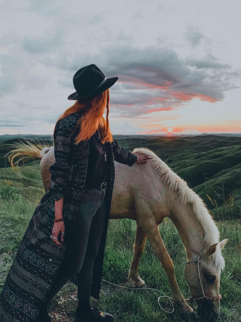 Puede incluir: Una mujer con un sombrero negro y un largo abrigo negro estampado se encuentra junto a un caballo blanco en un campo. El caballo est&aacute; pastando en la hierba. El cielo est&aacute; nublado con una puesta de sol en el fondo.