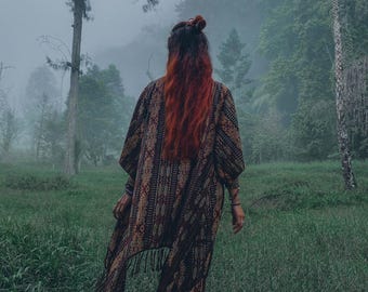 Puede incluir: Una mujer con el pelo largo y rojo camina por un bosque brumoso con un kimono largo y estampado con flecos. El kimono es marrón, negro y dorado con un diseño geométrico.