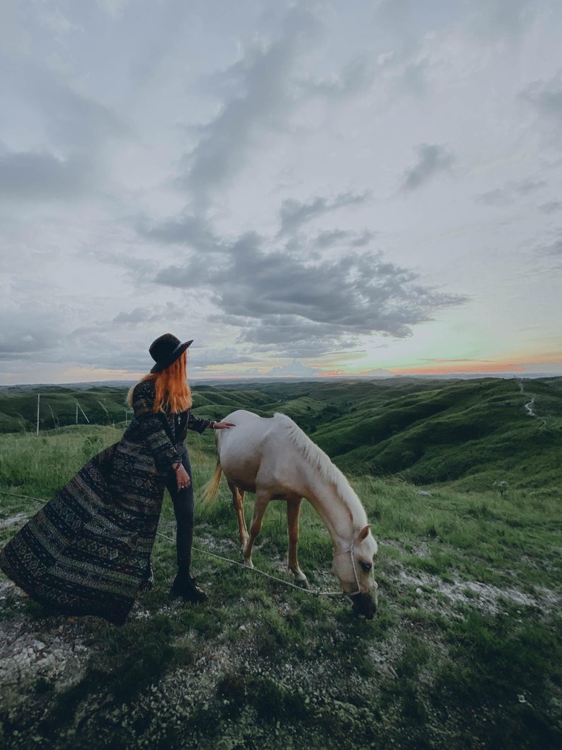Puede incluir: Una mujer con un abrigo largo estampado y un sombrero negro se encuentra en un campo con un caballo blanco. El caballo est&aacute; pastando en la hierba verde. El cielo est&aacute; nublado y el sol se est&aacute; poniendo en la distancia.
