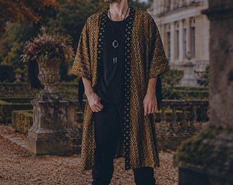 Puede incluir: Un kimono largo abierto por delante con un patrón geométrico de rombos en amarillo mostaza y marrón, con un borde contrastado de motivos en blanco y negro. La prenda se lleva sobre una camiseta negra lisa, pantalones negros y botas negras.