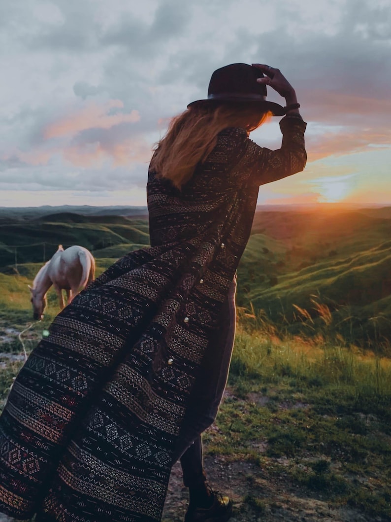 Puede incluir: Una mujer con un abrigo estampado en negro y marr&oacute;n con un sombrero negro de ala ancha se encuentra en una colina cubierta de hierba con un caballo blanco al fondo. El sol se pone en la distancia.