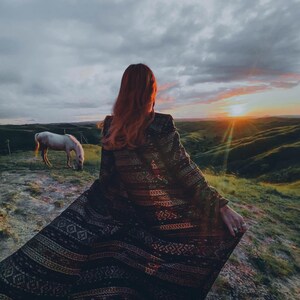 Puede incluir: Una mujer con el pelo largo y rojo lleva una chal de color negro y dorado con patrones y se encuentra de pie en la cima de una colina cubierta de hierba con vistas a una puesta de sol. Un caballo blanco pasta en primer plano.
