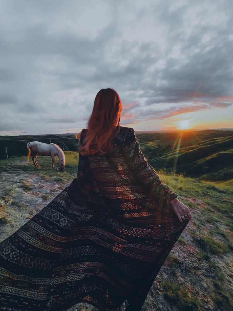 Puede incluir: Una mujer con el pelo largo y rojo lleva una chal de color negro y dorado con patrones y se encuentra de pie en la cima de una colina cubierta de hierba con vistas a una puesta de sol. Un caballo blanco pasta en primer plano.