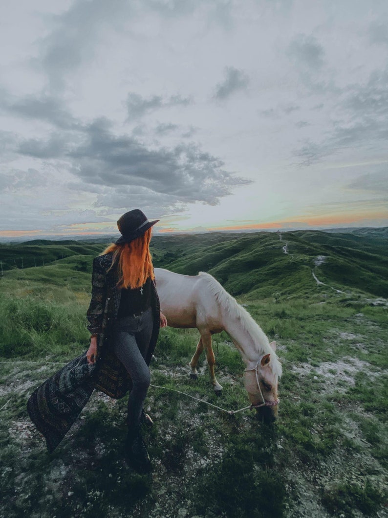 Puede incluir: Una mujer con un sombrero negro y un abrigo largo estampado se encuentra con un caballo blanco en un campo de hierba. El cielo est&aacute; nublado y el sol se pone en la distancia.