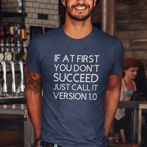 May include: A man wearing a navy blue t-shirt with the text "If at first you don't succeed just call it version 1.0" in white lettering. He is standing in a doorway with a wooden frame.