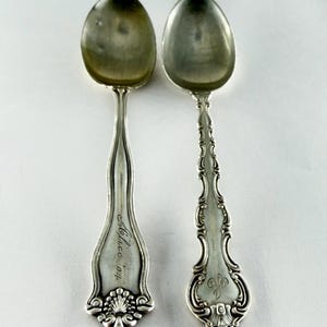 May include: Two antique silver spoons with ornate handles. One spoon is engraved with the name "Alice" and the initials "o.p." The other spoon has an ornate handle with a monogram. The spoons are displayed on a white surface.