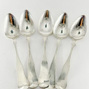 May include: Five silver spoons arranged on a white background. Each spoon has a rounded bowl and a long, slender handle. The spoons reflect the light, creating highlights and shadows. The spoons are likely for serving or eating.