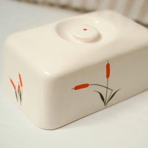 May include: Cream-colored rectangular ceramic butter dish with a small, round lid. The dish is decorated with orange cattail designs and black stems. The lid has a small, red dot in the center.