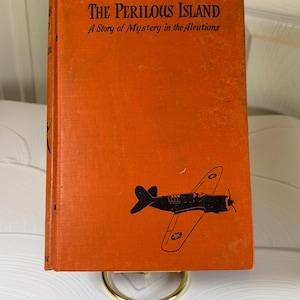 May include: An orange book titled "The Perilous Island: A Story of Mystery in the Aleutians" with a black and white illustration of a plane on the cover.