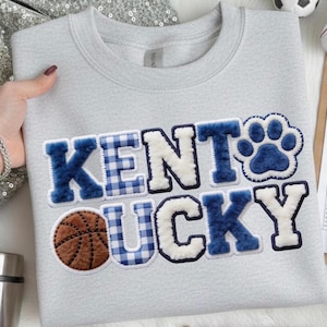 May include: A light gray sweatshirt with the words "KENTUCKY" in blue and white, with a basketball and paw print graphic. The letters are a mix of solid and plaid patterns. The sweatshirt is a crew neck style.
