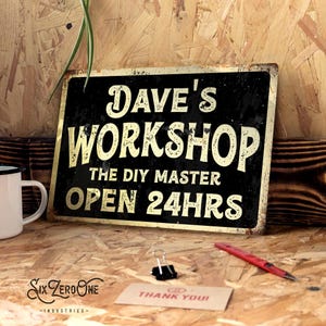 May include: A vintage-style black and gold metal sign that says "Dave's Workshop, The DIY Master, Open 24hrs." A white mug, a red pen, and a "Thank You!" card are on a wooden surface near the sign.
