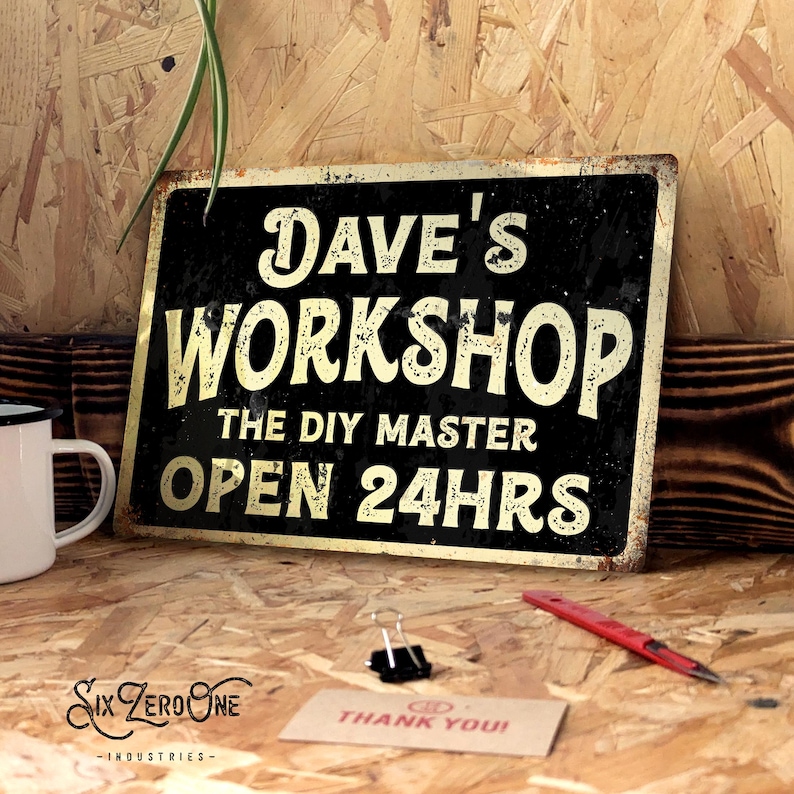 May include: A vintage-style black and gold metal sign that says "Dave's Workshop, The DIY Master, Open 24hrs." A white mug, a red pen, and a "Thank You!" card are on a wooden surface near the sign.