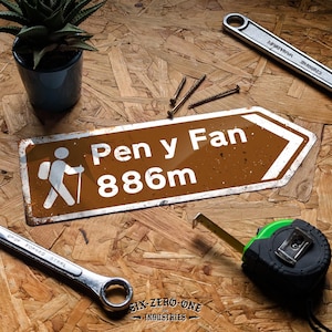 May include: A brown and white signpost reading "Pen y Fan 886m" with a hiker symbol, alongside tools like a wrench, tape measure, screws, and a plant in a pot. The sign is set on a wooden surface. The height is 886 meters.