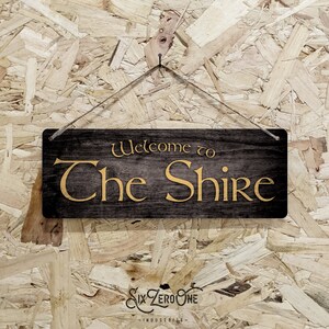 May include: A wooden sign with the text "Welcome to The Shire" in gold lettering on a black background. The sign is hanging from a string against a wood chip background.