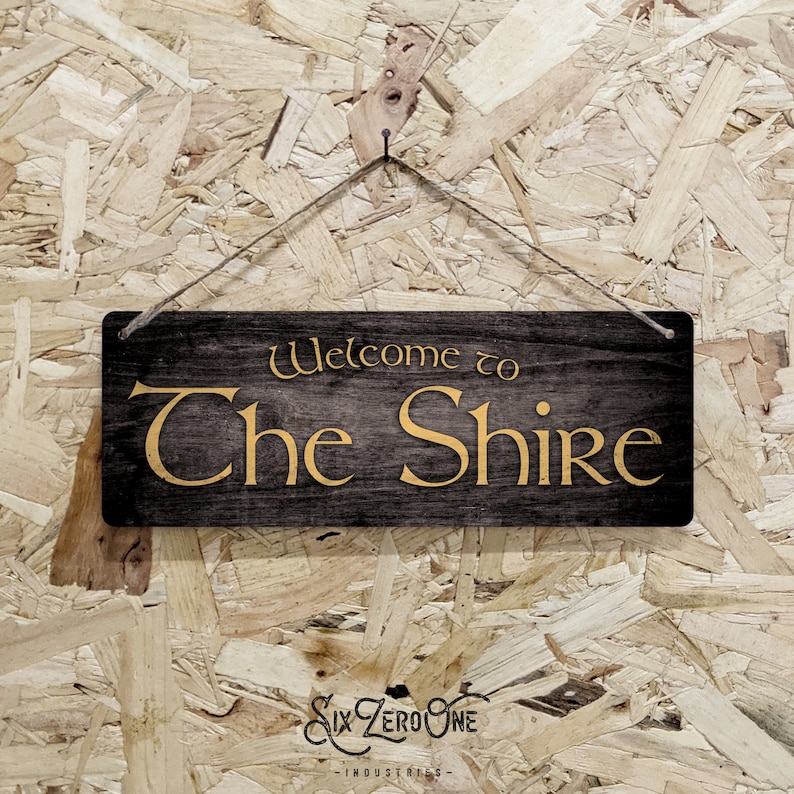 May include: A wooden sign with the text "Welcome to The Shire" in gold lettering on a black background. The sign is hanging from a string against a wood chip background.