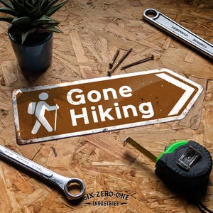 May include: A brown and white "Gone Hiking" sign featuring a hiker graphic, alongside tools such as a wrench, tape measure, and screws. A small potted plant is also visible. The sign's message suggests a theme of outdoor pursuits and DIY projects.