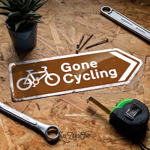 May include: A brown sign with the text "Gone Cycling" and a bicycle illustration. The sign is surrounded by tools, including a spanner, tape measure, and screws, on a wood-grain surface. A small potted plant is also present.