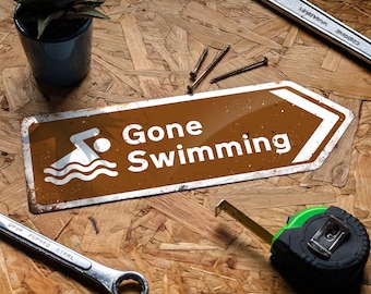 May include: A brown and white "Gone Swimming" sign with a swimmer graphic, surrounded by tools like a wrench, tape measure, and screws. A small potted plant is also present. The sign is on a wood-grain surface.
