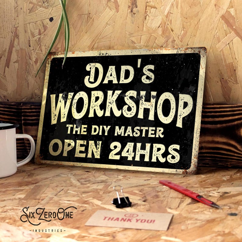 May include: A black metal sign with gold lettering that reads "DAD'S WORKSHOP THE DIY MASTER OPEN 24HRS." The sign has a distressed, rustic appearance. A white mug, a red pen, and a "THANK YOU!" card are on the wooden surface.