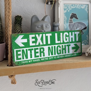 May include: A weathered green metal sign with white lettering, reading "EXIT LIGHT ENTER NIGHT". The sign also features the text "TAKE MY HAND, WE'RE OFF TO NEVER-NEVER LAND". A grey cat head statue is positioned above the sign.