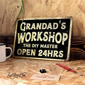 May include: A black and gold metal sign with the text "Grandad's Workshop, The DIY Master, Open 24hrs." The sign has a distressed, vintage appearance. A white mug, a red pen, and a "Thank You!" card are also present.